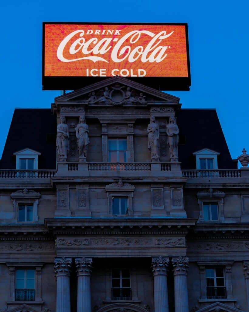 Advertising. Illuminated Coca-Cola billboard atop an ornate historic building against a twilight sky.